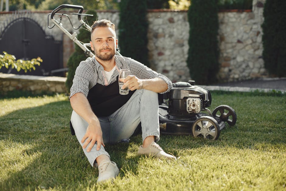 A man with a neatly trimmed beard and short hair, wearing a checkered shirt and an apron, sits on a well-maintained lawn in a garden, holding a glass of water and smiling at the camera. Behind him, th